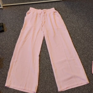 Ljusrosa wide byxor från Elitzia - Säljer ett par ljusrosa byxor från Elitzia i storlek S. De har en skön, bred passform med resår i midjan och är tillverkade i ett mjukt bomullsmaterial. Perfekta för chill dagar eller när du vill ha en avslappnad look. Kan tvättas innan jag skickar det om du vill😄