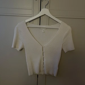 Vit ribbad topp med knappar från H&M - Säljer en vit croppad topp från H&M i storlek XS. Toppen har ribbad struktur, korta ärmar och v-ringad hals med knappar framtill. Perfekt för dig som gillar enkel och stilren design med en liten twist. Superfin att matcha med jeans eller kjol!