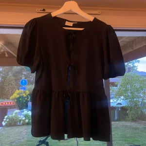 Svart blus med knyt från Gina Tricot - Supersöt svart blus från Gina Tricot med puffiga korta ärmar och volang nedtill. Framsidan har två knytband som ger en extra detalj. Perfekt för dig som gillar en feminin och trendig look. Tunt och lätt material som känns skönt mot huden. Aldrig använt!