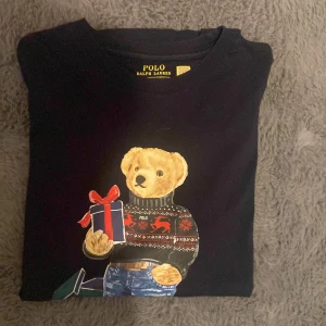 Blå Polo Bear t-shirt från Ralph Lauren - Mörkblå t-shirt från Polo Ralph Lauren med ikonisk Polo Bear-tryck framtill. Björnen har jultröja, jeans och håller en present med röd rosett. 