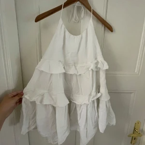 Vit volangklänning med halterneck - Superfin vit kortklänning med volanger och halterneck. Klänningen har en luftig passform och är ärmlös med tunna knytband i nacken. Perfekt för varma dagar när du vill känna dig både söt och bekväm.