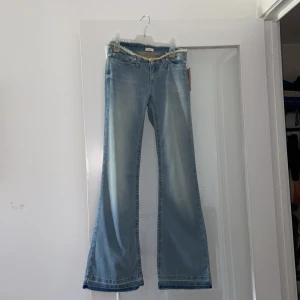 Ljusblå bootcut jeans från Nelly - Snygga ljusblå jeans från Nelly med bootcut-modell och fransad midja. Jeansen har klassisk femficksdesign, gylf med dragkedja och knäppning samt coola slitningar vid midjan. Perfekta för dig som gillar en retro vibe och vill sticka ut lite extra.