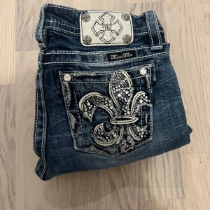 Miss Me jeans med broderade bakfickor - Snygga blå Miss Me jeans med bootcut och coola broderade bakfickor med stora fleur-de-lis och blingdetaljer. Jeansen har slitningar och kontrastsömmar, samt en unik patch med metallknappar i midjan. Perfekta för dig som gillar statement-jeans! Längd 95cm midja 33-34 benöppning 21 inseam 73