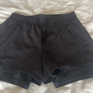Svarta träningsshorts  - Svarta träningsshorts från H&M Move med elastisk midja och snörning. Shortsen har en loose passform och är tillverkade i ett lätt syntetmaterial som andas, perfekt för sport eller gym. Snygg och enkel design utan mönster, med diskreta meshpaneler på sidorna. Dem är supertunna och sköna att träna i och passar perfekt en varm sommardag. Passar en xs. 