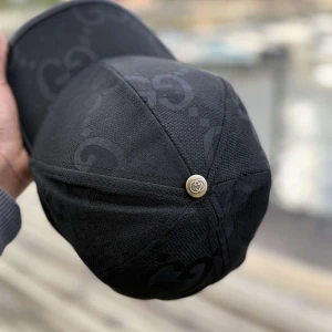 Svart keps från Gucci med GG-mönster - Snygg svart keps från Gucci med diskret GG-monogram över hela kepsen. Klassisk böjd skärm med svart kant och metallknapp med Gucci-logo på toppen. Tillverkad i bomull och polyester för en lyxig känsla och perfekt passform.