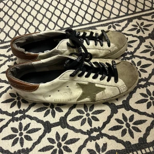 Golden Goose vita sneakers med stjärna - Golden Goose sneakers i vitt skinn med olivgrön stjärndetalj på sidan och bruna detaljer vid hälen. Svarta snören med logga och rund tå. Original sulan är inte kvar
