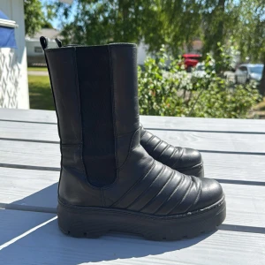 Svarta chunky boots med platå - Chunky svarta boots i skinnimitation med högt skaft och bred resår på sidorna. Snygg quiltad design framtill och kraftig platåsula med grovt mönster undertill. Perfekta för dig som gillar en edgy och modern look.