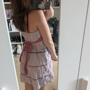 Randig sommarklänning från Mango - Söt kortklänning från Mango med rosa, blå och vit randigt mönster. Klänningen har v-ringning, knyt i ryggen och smala axelband. Tillverkad i bomull, perfekt för varma dagar. Lätt och luftig, perfekt till sommaren. 