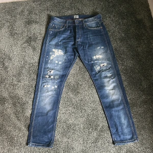 Blå slitna jeans från Jack & Jones - Säljer ett par blå jeans från Jack & Jones, modell Erik Original Vintage. Jeansen har slitna detaljer och hål framtill för en cool, avslappnad look. Klassisk femficksmodell med raka ben och normal passform. Perfekta för dig som gillar streetstyle.