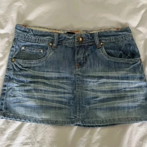 Blå jeanskjol med fickor - Snygg blå jeanskjol från Lindex med klassisk femficksdesign, bälteshällor och slitna detaljer framtill. Kjolen är kort och har en rak passform, perfekt till sneakers eller boots. Stängs med knapp och dragkedja framtill.