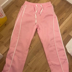 Rosa mjukisbyxor med vita ränder - Säljer ett par ljusrosa mjukisbyxor med vita ränder längs sidorna och snörning i midjan. Byxorna har en loose passform och är perfekta för en chill och avslappnad stil. Resår i midjan för extra komfort.