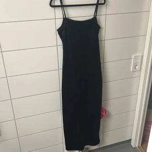 Svart lång sammetsklänning - En stilren svart långklänning i mjuk sammet med smala axelband och rak passform. Perfekt för dig som gillar minimalistisk och elegant stil. Klänningen har en enkel design utan detaljer och är lätt att styla med accessoarer.