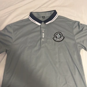 Grå Moncler pikétröja med logga - Snygg grå pikétröja från Moncler med klassisk krage i mörkblått och vitt. Tröjan har korta ärmar och tydlig Moncler-logga på bröstet. Perfekt för dig som gillar stilrena och sportiga vibes.