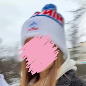Vit Minttu mössa med blå tofs - Snygg vit mössa från Minttu med blå och röda detaljer, blå ränder och stor blå-vit tofs på toppen. Broderad Minttu-logga framtill. Perfekt för kalla dagar och ger en cool retrovibe.