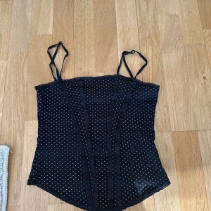 Svart prickig korsett från H&M XS - Supersöt svart korsett-topp från H&M i storlek XS. Tunt tyg med små vita prickar, smala axelband och spetsdetaljer framtill. Figurnära passform och lätt croppad längd. Perfekt för att lyfta vilken outfit som helst.