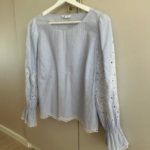 Blåvit blus med spetsdetaljer - Supersöt blus i ljusblått och vitt med tunna ränder, broderade spetsdetaljer längs ärmar och nederkant. Perfekt för vår och sommar. Storlek 36 och knappt använd. 🩷🩷