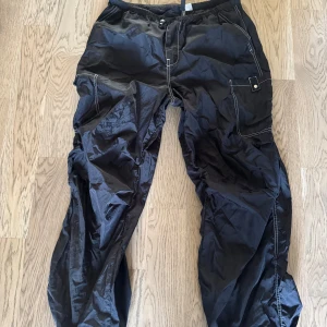 Svarta cargopants från H&M Divided - Svarta cargopants från H&M Divided i storlek S. Byxorna har flera fickor, resår i midjan och synliga kontrastsömmar. Materialet är lätt och syntetiskt, perfekt för en avslappnad streetstil.