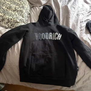 Hoodrich hoodie - Svart hoodie från Hoodrich med stor broderad logga i vitt och grått på bröstet. Klassisk känguruficka framtill, justerbar huva med snören och mjukt material. Perfekt för en chill och street-inspirerad look.
