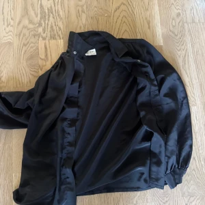 Svart satinliknande skjorta New Editions - Snygg svart skjorta från New Editions i glansigt, satinliknande polyestertyg. Klassisk krage, långa ärmar med knäppning vid ärmslut och knappar framtill. Perfekt för dig som gillar en stilren och elegant look.