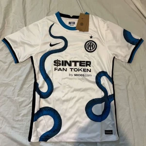 Inter Milan bortatröja 2021/22 Nike - Inter Milans vita bortatröja från säsongen 2021/22 med blå ormmönster, svarta detaljer och klubbmärke på bröstet. Tröjan har korta ärmar, blå och svarta muddar samt tryck från sponsorer. Tillverkad i lätt och ventilerande polyester.