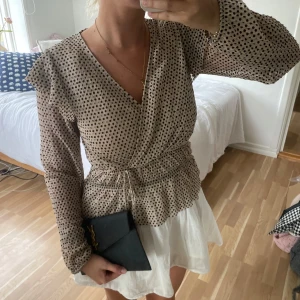 Beige prickig omlottblus med knyt🤎 - Supersöt beige omlottblus med svarta prickar och knyt i midjan. Långärmad modell med lätt volang vid axlarna och v-ringning. Tunt, luftigt material som känns skönt mot huden och ger en avslappnad men chic vibe🤎