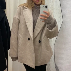 Beige oversized kappa med knappar - Snygg beige kappa i oversized modell med stora slag och svarta knappar framtill. Jackan har en rak, boxig passform och är perfekt för lager-på-lager. Stilren och enkel att matcha till olika outfits.