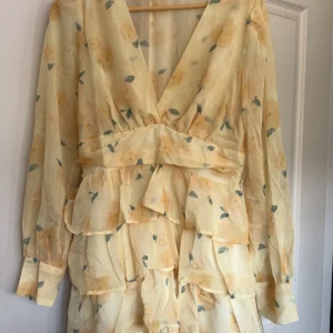 Ljusgul blommig volangklänning - Superfin ljusgul kortklänning med blommigt mönster och volanger. Djup v-ringning, långa ärmar med knapp vid mudden och omlottkänsla i midjan. Tunt, lätt material som känns luftigt och perfekt för varma dagar.