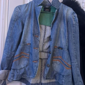 Blå jeansjacka från Lee XS - Snygg blå jeansjacka från Lee i klassisk modell med coola orangea sömmar och detaljer. Jackan har två fickor framtill, knappar och en figursydd passform. Perfekt för dig som gillar en avslappnad men ändå stilren look.