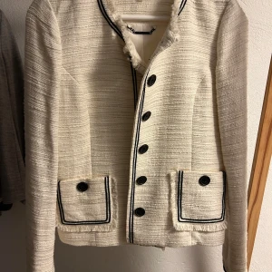 Vit kavaj från Karl Lagerfeld Paris - Snygg vit kavaj från Karl Lagerfeld Paris med svarta knappar och svarta detaljer längs kanterna. Jackan har två fickor framtill med dekorativa knappar och fransade kanter vid kragen. Tillverkad i ett strukturerat tyg som ger en exklusiv känsla.