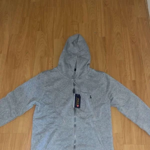 Grå kofta - Snygg grå hoddie dragkedja och huva. Klassisk logga broderad på bröstet och praktiska fickor framtill. 