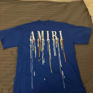 Blå AMIRI t-shirt med färgstänk - Snygg blå t-shirt från AMIRI med coolt färgstänksmönster och stor logga på ryggen. Framsidan har en mindre logga med samma färgstänk. Klassisk passform och rund hals. Perfekt för dig som vill sticka ut med streetstyle-vibe. Storlek: M, stor i storlek passar L och kanske XL