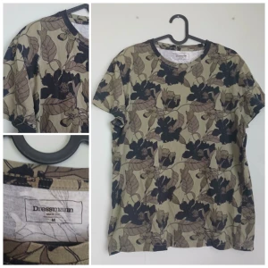  t-shirt från Dressmann M - Snygg t-shirt från Dressmann i storlek M med coolt blommönster i svart och brunt på en beige bakgrund. Klassisk rund halsringning och korta ärmar. Perfekt för dig som vill sticka ut lite extra med en unik print. Gjord i mjuk bomull för skön känsla.