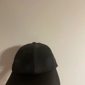 Svart keps från Prada i nylon - Snygg svart keps från Prada med klassisk böjd skärm och justerbart spänne bak. Kepsen har Pradas ikoniska metallogo på sidan och är tillverkad i glansig nylon som ger en lyxig känsla. Perfekt accessoar för en clean och stilren look.