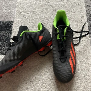Adidas svarta fotbollsskor strl 41⅓ - Snygga svarta fotbollsskor från Adidas med röda detaljer och neongrön insida. Skorna har snörning, mönstrad ovandel och röda dobbar för bra grepp på planen. Perfekta för dig som vill sticka ut med stil på matchen.