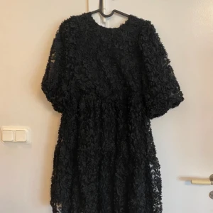 Svart kortklänning från Gina Tricot - Supersnygg svart kortklänning från Gina Tricot med 3D-blommig struktur och transparent mesh. Puffiga korta ärmar och snygg snörning i ryggen ger en unik look. Perfekt för dig som vill sticka ut med coola detaljer och trendig design.