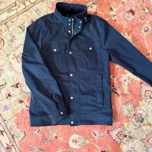 Marinblå vindjacka från J.Lindeberg - Snygg marinblå vindjacka från J.Lindeberg med hög krage, silverfärgade knappar och dragkedja. Jackan har fyra fickor framtill och diskret logodetalj på armen. Perfekt för dig som gillar stilren och sportig look.