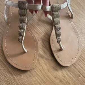 Beiga sandaler med glittriga detaljer - Snygga beiga sandaler med platt sula och T-rem dekorerad med glittrande stenar. Remmarna är i syntetmaterial och sandalerna har öppen tå. Perfekta för sommardagar när du vill ha något stilrent men ändå extra