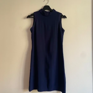 Marinblå ärmlös klänning Massimo Dutti - Stilren marinblå klänning från Massimo Dutti med hög krage och ärmlös design. Klänningen är kort och har en rak passform, perfekt för en clean och modern look. 