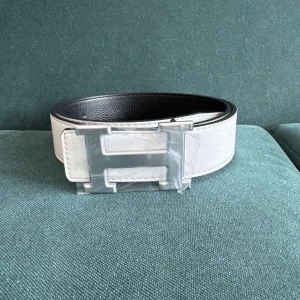 vitt skärp med silver H-spänne - Snyggt vitt bälte från med ikoniskt H-spänne i silver. Bältet är gjort i skinn och har en stilren, rak design med svarta kanter och insida. Perfekt accessoar för att lyfta din outfit med en lyxig touch.
