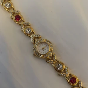 Guldig klocka med strass och fina stenar - Säljer en guldig super fin vintage armbandsklocka från Parmex quanta som inte längre existerar, den har glittrande strass runt urtavlan och på länken. Klockan har vita, röda och gröna stenar i armbandet och en rund vit urtavla med små kristaller som timmarkeringar, Perfekt för dig som gillar bling och unika detaljer ❣️
