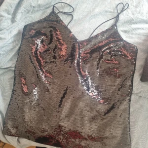 Glittrig paljettopp med tunna band - Supersnygg glittrig topp med tunna axelband och v-ringning. Toppen är täckt av skimrande paljetter i svart som skimrar som silver och bronsiga toner, perfekt för att sticka ut. Insidan är fodrad med mjuk satin för extra komfort. Passar dig som vill ha en statement-piece till festen! Liten slits i sidan för att det ska sitta snyggt!