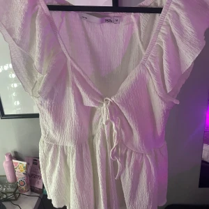 Vit volangblus med knyt från 157 - Supersöt vit blus från 157 med volangdetaljer längs ärmarna och v-ringning. Blusen har knyt framtill och är tillverkad i ett lätt, skrynkligt bomullstyg. Perfekt för dig som gillar romantisk och luftig stil.