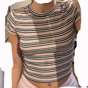 Randig croppad topp från H&M - Säljer en croppad, ribbad topp från H&M i ljusrosa med svarta och vita ränder. T-shirten har korta ärmar och rund halsringning. Tillverkad i ekologisk bomull och sitter tight mot kroppen. Perfekt för dig som gillar en enkel men snygg stil. Passar för barn också. Använd minst 3 gånger. 