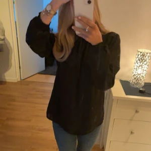 Svart blus med smockad krage - Snygg svart blus med smockad hög krage och långa ärmar. Stängning med knapp i nacken och diskreta puffärmar ger en trendig look. 🌟