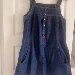 Jeansklänning med knappar från Ellos - Snygg jeansklänning från Ellos Plus Collection i 100% bomull. Mörkblå denim, breda axelband med spännen och knappar framtill. Praktiska fickor och kontrastsömmar ger en cool vibe. Perfekt för dig som gillar avslappnad stil med edge.