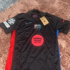 FC Barcelona Pedri #8 svart matchtröja - Snygg svart FC Barcelona fotbollströja från Nike med Pedri och nummer 8 på ryggen. Tröjan har röda och blå detaljer på sidorna, Champions League-märke på ärmen och stora röda tryck med Spotify-logga framtill. Tillverkad i lätt och ventilerande material.