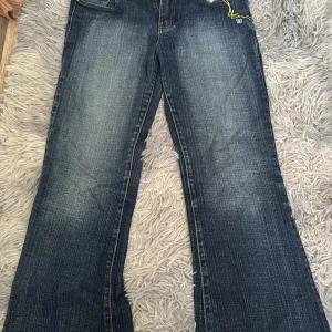 Gallop bootcut jeans med brodyr - Mörkblå bootcut jeans från Gallop med snyggt slitna detaljer och kontrasterande gula sömmar. Fickor med broderade mönster och liten broderad fjäril både fram och bak. Klassisk femficksmodell i jeansmaterial.