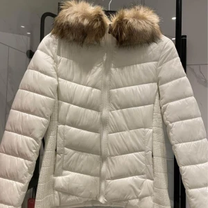 Vit pufferjacka med fuskpäls från Etirel - Snygg vit pufferjacka från Etirel med fluffig fuskpäls runt huvan. Jackan har quiltad design, dragkedja framtill och ribbade partier på sidorna. Perfekt för kalla dagar när du vill hålla stilen och värmen.