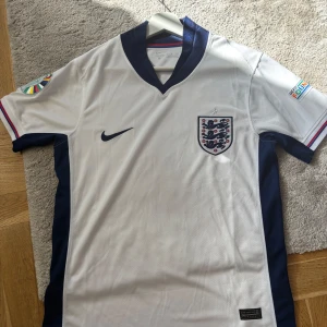 England Nike fotbollströja vit/blå S - England fotbollströja från Nike i vitt med marinblå detaljer på sidor och krage. Broderat landslagsmärke på bröstet, Nike-logga och UEFA Foundation-märke på ärmen. Tillverkad i lätt Dri-FIT polyester som andas. Klassisk passform med korta ärmar.