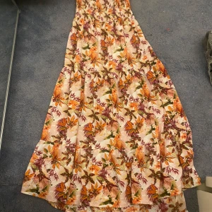 Blommig långklänning i somriga färger storlek 158 ( barn) - Superfin långklänning med blommigt mönster i orange, lila och grönt på ljus botten. Klänningen har smock upptill och volangig kjol, perfekt för varma dagar. Luftigt material och riktigt skön passform. 